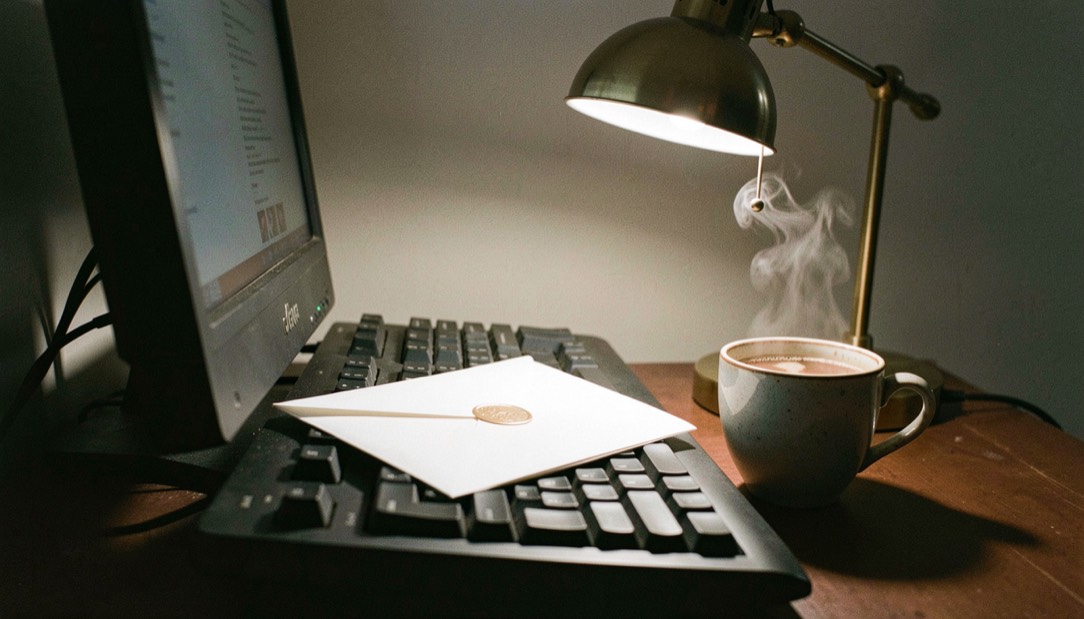 Une enveloppe blanche posée sur un clavier d'ordinateur à côté d'une tasse de café fumante, éclairée par une lampe de bureau en laiton dans une pièce sombre